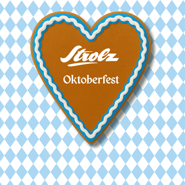 Ein herzförmiger Lebkuchen mit dem Strolz-Oktoberfest-Logo, verziert mit blau-weißer Zuckerglasur. Im Hintergrund ist ein traditionelles bayerisches Rautenmuster in Blau und Weiß zu sehen. Das Motiv vermittelt eine festliche, gemütliche Oktoberfest-Atmosphäre.